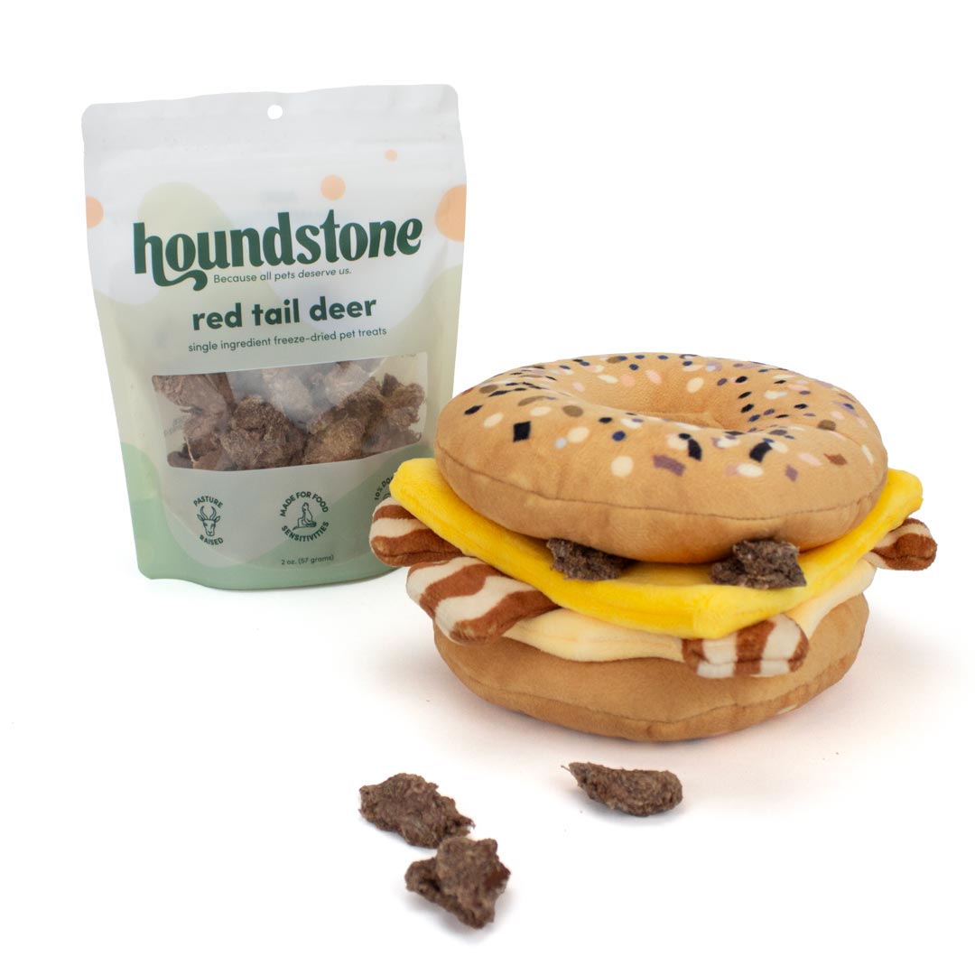 Houndstone Red Tail Deer single ingredient freeze-dried cat treats shown next to a plush toy shaped like a bagel cheese burger, with a few loose cat treats in front on a white background.