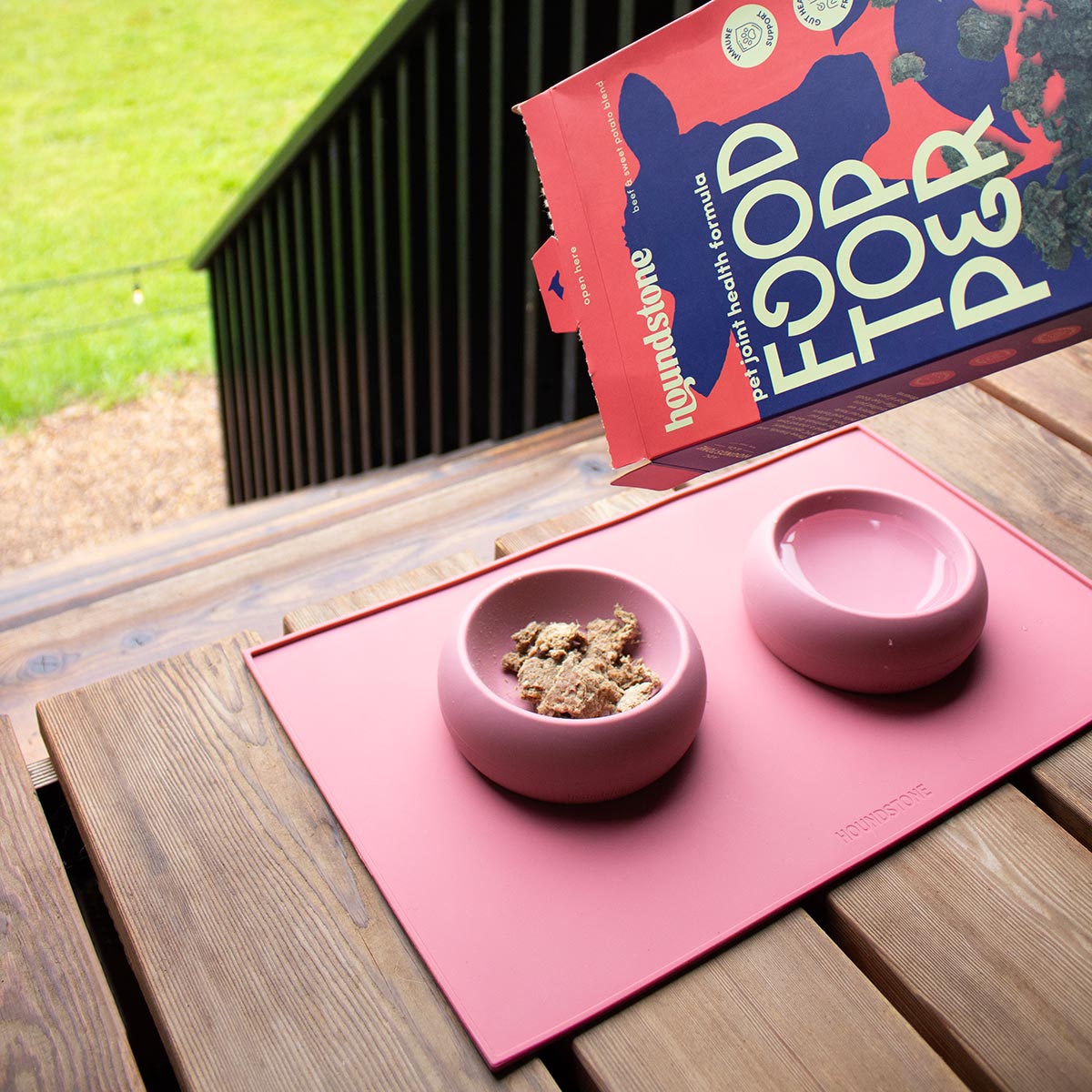 Pink dog bowls with food on a pink dog food mat, with a dog food topper package in the background.