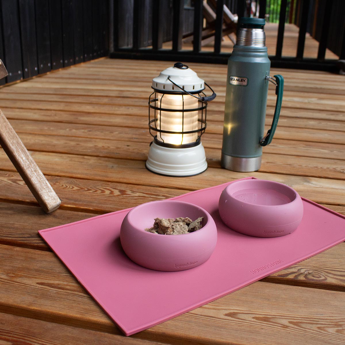 Pink dog bowls with food on a pink dog food mat on a wooden deck with a lantern and thermos in the background.