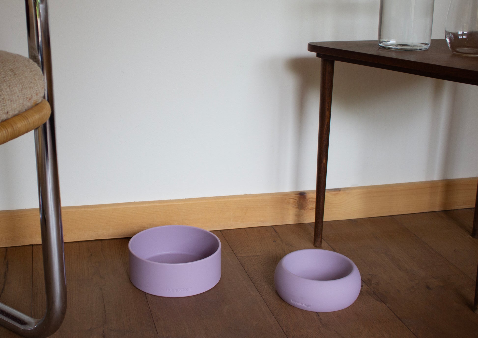 Two purple dog bowls on a wooden floor with a white wall and table in the background.