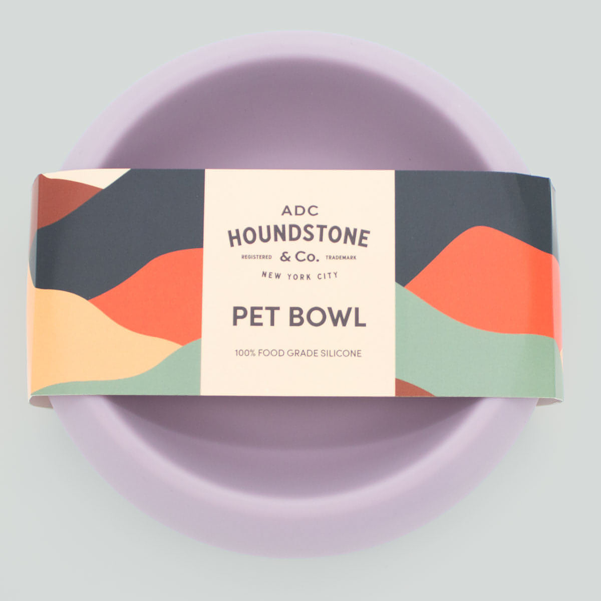 A silicone dog bowl in mauve color with a non-slip base packaging that has multicolored waves and the brand 'Houndstone & Co.' printed on it.